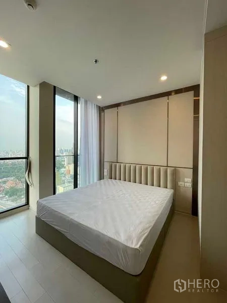 Condominium for rent in Pathum Wan, Bangkok - Master bedroom with a large bed and corner floor-to-ceiling windows overlooking the city.