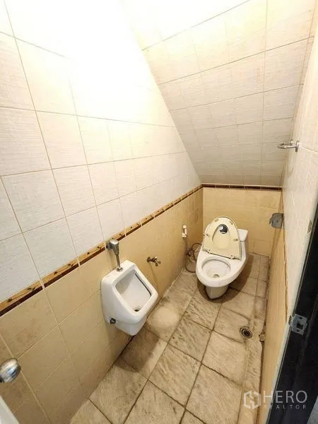 Shophouse for rent in Bang Rak, Bangkok - Compact tiled restroom with sloped ceiling, toilet and wall urinal.