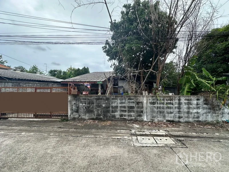 地 出售在Phra Khanong, Bangkok - 正面宽幅视图：低墙、树后房屋与沿街架空电线。