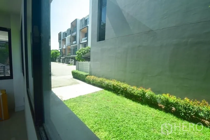 Townhouse for rent or sale in Suan Luang, Bangkok - View from a window to a private lawn and a neat row of townhouses on a sunny day.