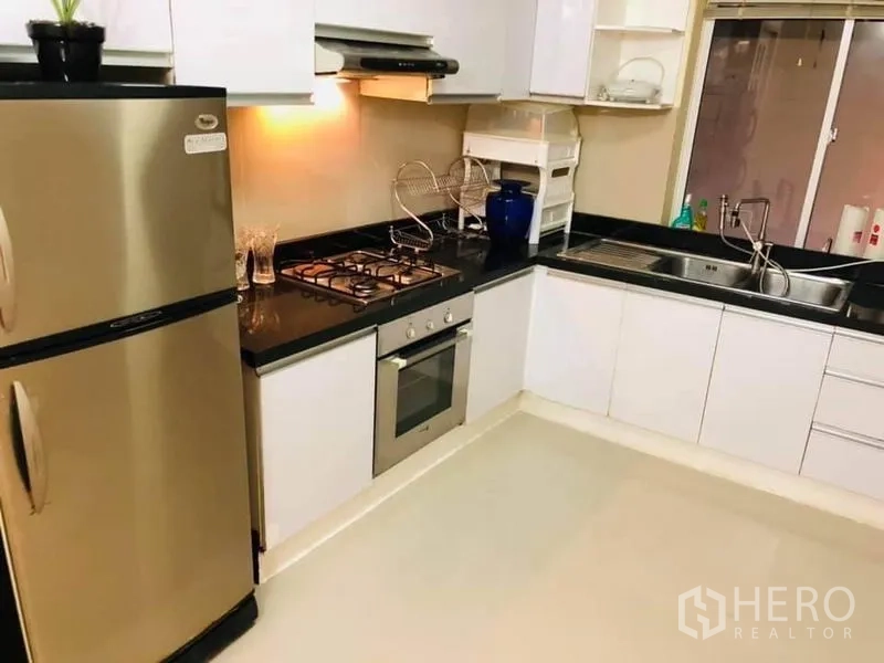 Detached House for rent in Watthana, Bangkok - Modern L-shaped kitchen with black granite countertop, gas hob, oven, and stainless steel refrigerator.