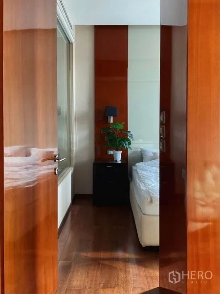 Condominium for rent in Khlong Toei, Bangkok - Entrance view into bedroom with warm wood panels, bedside table, and potted plant.