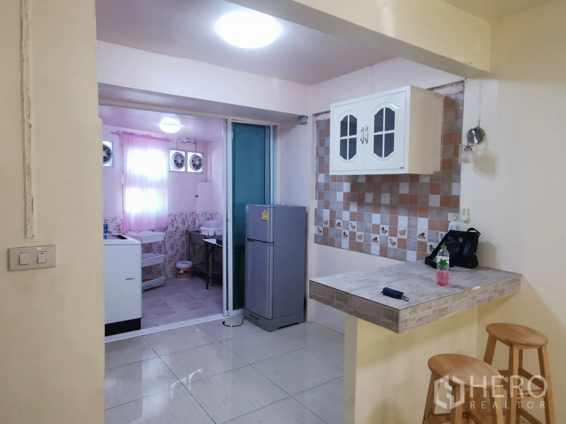 Townhouse for rent in Khlong Toei, Bangkok - Kitchen corner with checkered backsplash, hanging cabinet, breakfast counter, and sliding glass to the utility area.