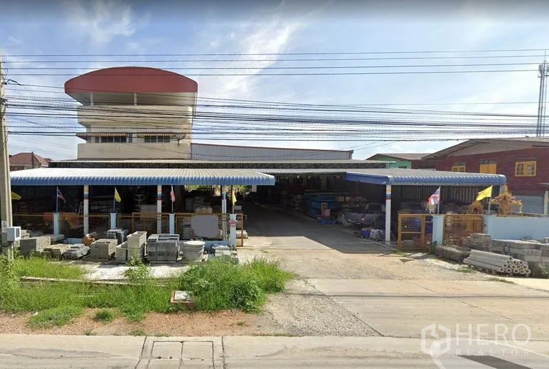 Warehouse for rent in Sai Noi, Nonthaburi - Street-facing exterior of a Sai Noi warehouse with blue metal canopies, wide driveway and stacked building materials.