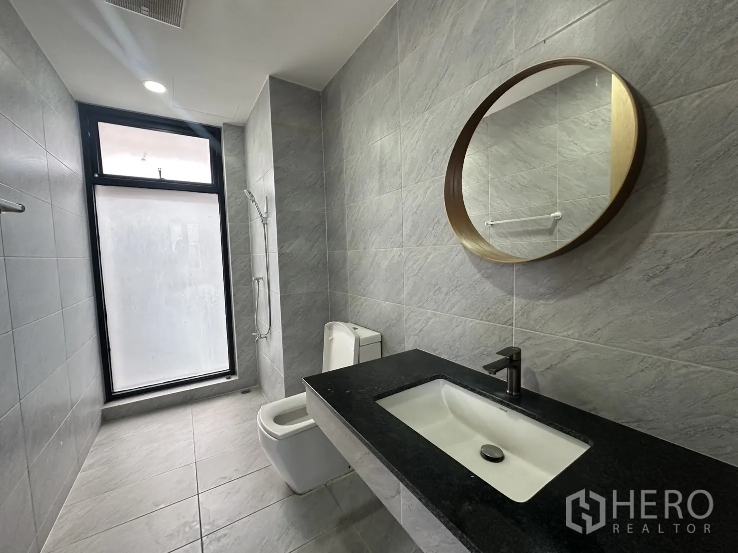 Home Office for rent in Pathum Wan, Bangkok - Gray-tiled bathroom with round mirror, vanity and toilet beside a frosted window.