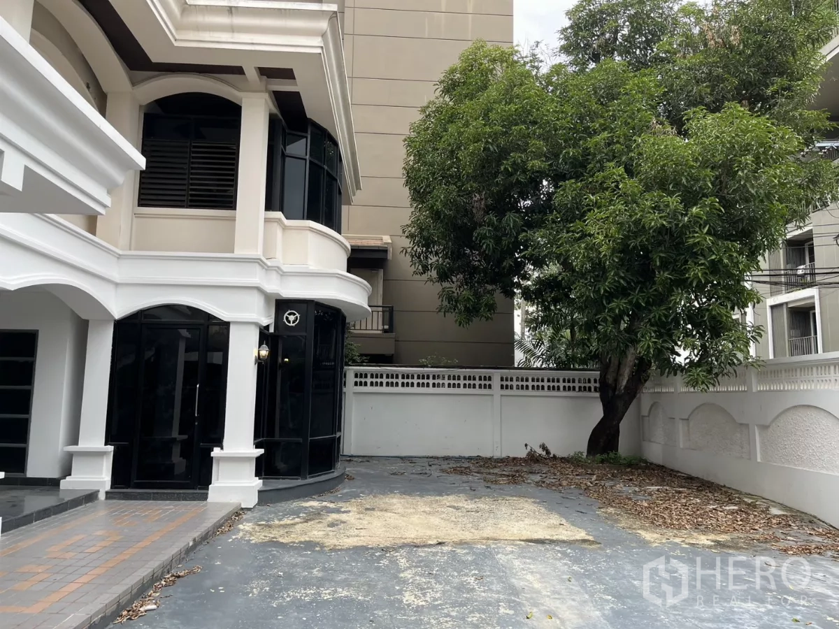 独立式住宅 出租在Watthana, Bangkok - 侧面外观，转角落地窗、宽阔车道与一棵大树。