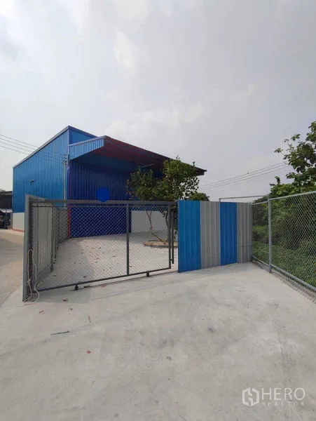 Warehouse for rent in Pak Kret, Nonthaburi - Gated entrance with chain-link fence leading to the warehouse yard and parking area.