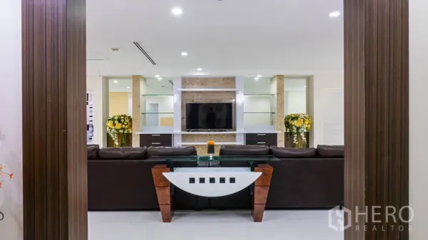 Condominium for rent or sale in Khlong Toei, Bangkok - Living room with dark leather sofa facing a wall-mounted TV and built-in shelving.