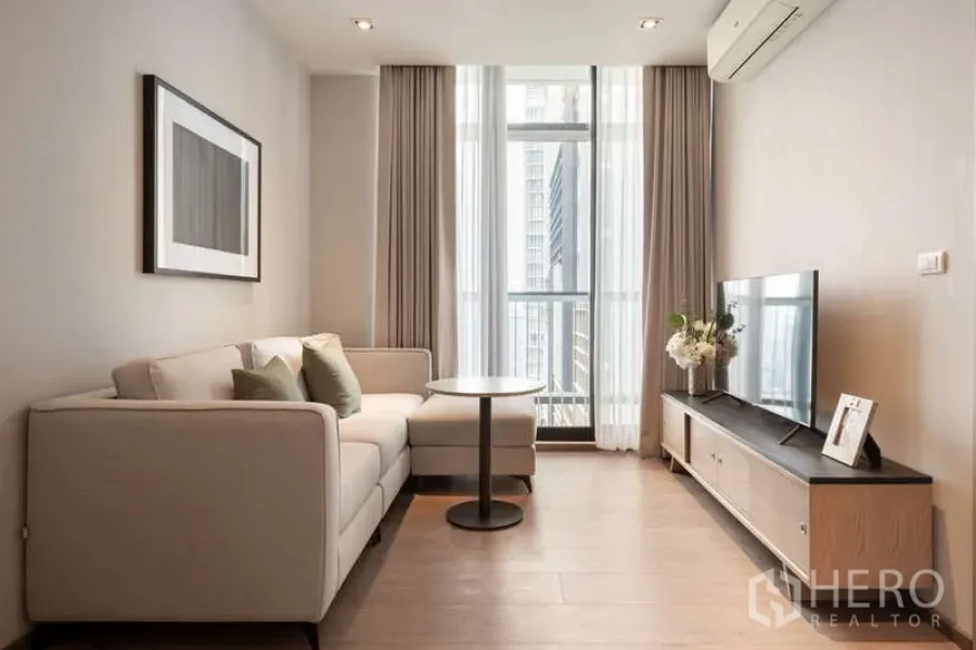 Condominium for rent in Khlong Toei, Bangkok - Living area with sofa and TV facing tall windows and balcony, modern neutral decor.