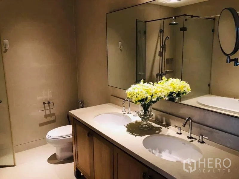 Condominium for rent in Khlong Toei, Bangkok - Spacious bathroom with double vanity, large mirror, glass shower and toilet in a Bangkok condo.