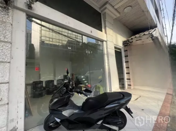 零售空间 出租 - 玻璃门店入口的斜角视图，旁有瓷砖人行道与停放的踏板车。