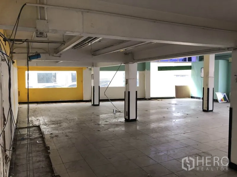 Warehouse for rent in Suan Luang, Bangkok - Spacious ground-floor warehouse area with pillars, tiled floor, exposed wiring for lighting, and bright street-facing windows.
