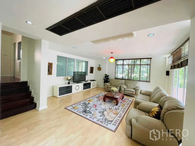 Detached House for rent in Watthana, Bangkok - Spacious lounge with TV console, stairs, and corner windows overlooking greenery.