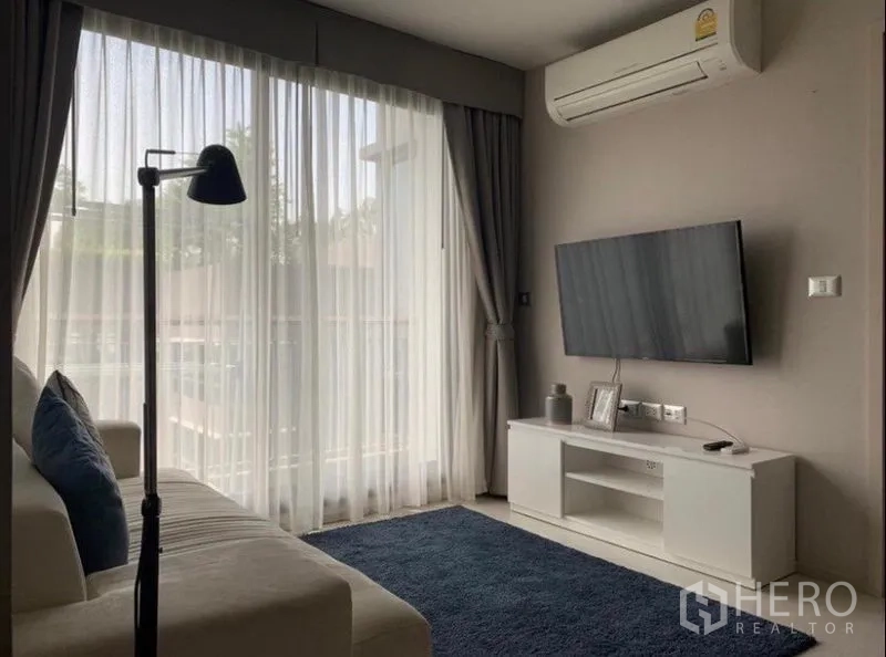 Condominium for rent in Khlong Toei, Bangkok - Living area with wall-mounted TV, blue rug, and sliding glass doors to a balcony with sheer drapes.