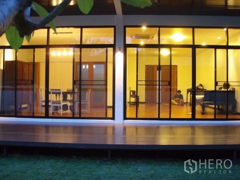 Detached House for rent in Huai Khwang, Bangkok - Evening view of modern single-storey house with full-height sliding glass walls, wood deck and lawn