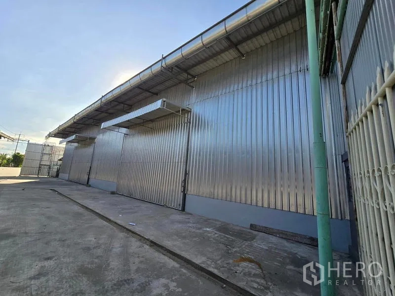 Warehouse for rent in Sai Noi, Nonthaburi - Side elevation of corrugated metal warehouse with multiple service doors, roof guttering, and a paved yard.