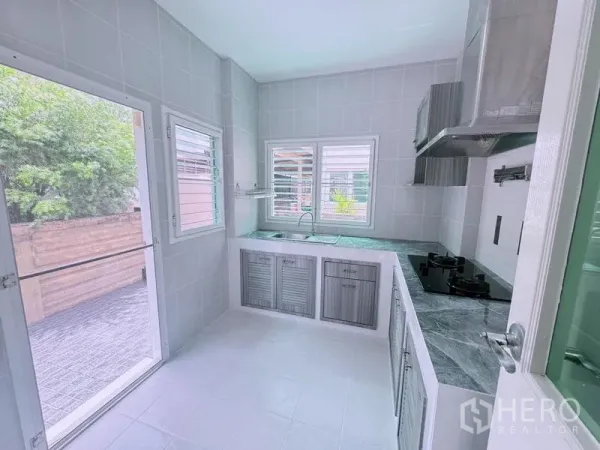 Detached House for rent in Prawet, Bangkok - Bright tiled kitchen with L-shaped counters, gas hob, stainless hood, and windows opening to the patio.