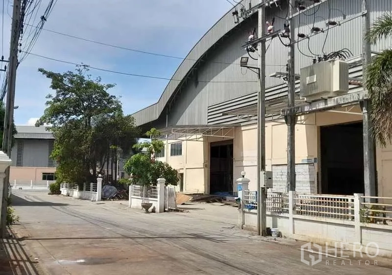 Factory for rent in Mueang Samut Prakan, Samut Prakan - Exterior driveway beside the factory with roll‑up doors, fencing and utility poles.