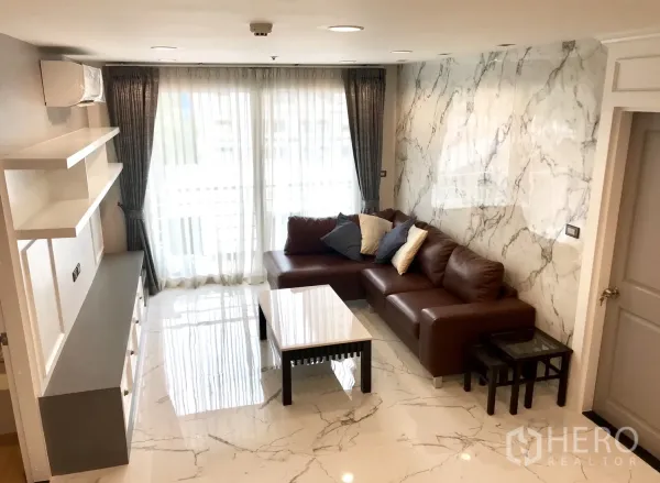 Condominium for rent or sale in Khlong Toei, Bangkok - Bright living room with marble-effect floors, L-shaped brown leather sofa and large sheer-curtained windows.