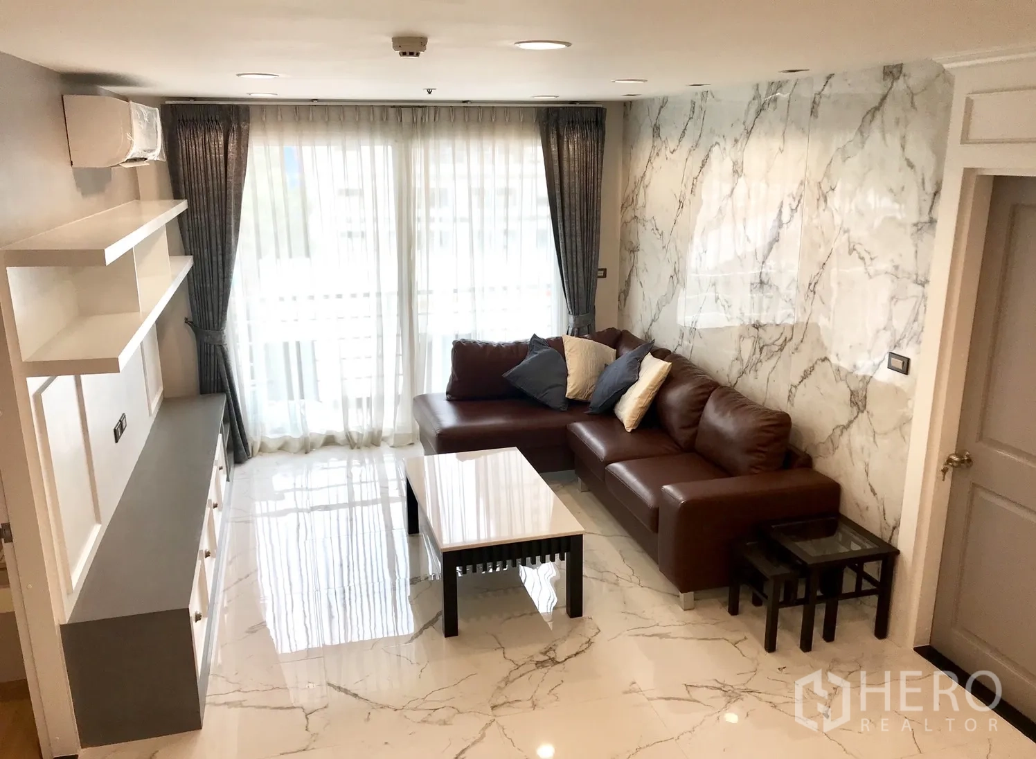 Condominium for rent or sale in Khlong Toei, Bangkok - Bright living room with marble-effect floors, L-shaped brown leather sofa and large sheer-curtained windows.