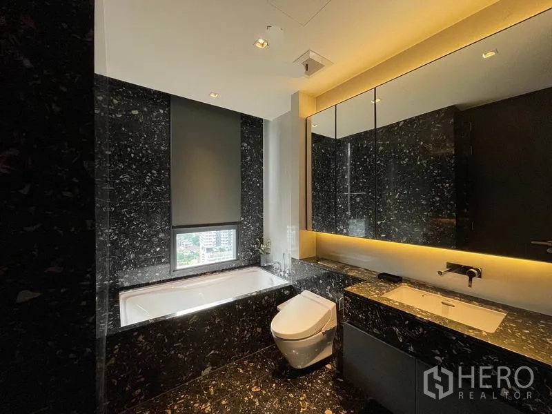 Condominium for rent in Khlong Toei, Bangkok - Luxurious bathroom with dark marble finishes, bathtub, backlit mirror and modern fixtures.