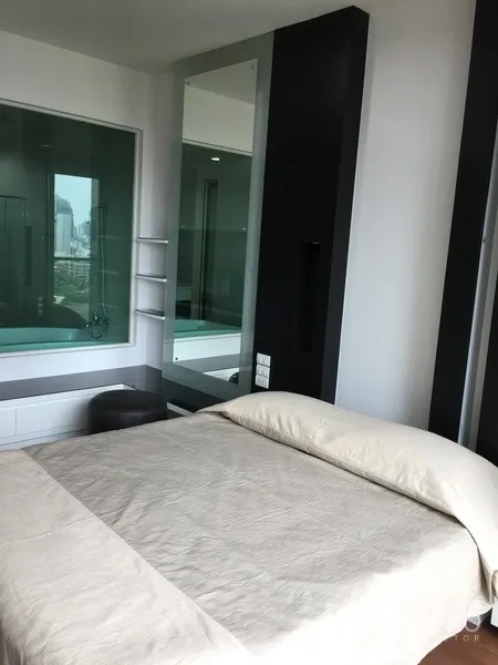 Condominium for rent in Pathum Wan, Bangkok - Bright bedroom with beige bedding, glass window to bathroom featuring a bathtub and a distant city view.