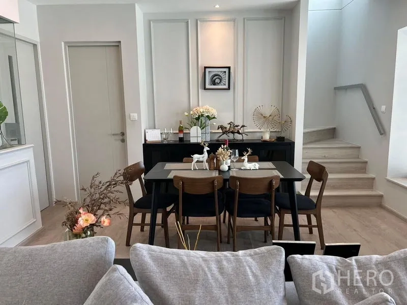 Detached House for rent or sale in Prawet, Bangkok - Dining area for six beside a sideboard and staircase with soft lighting.