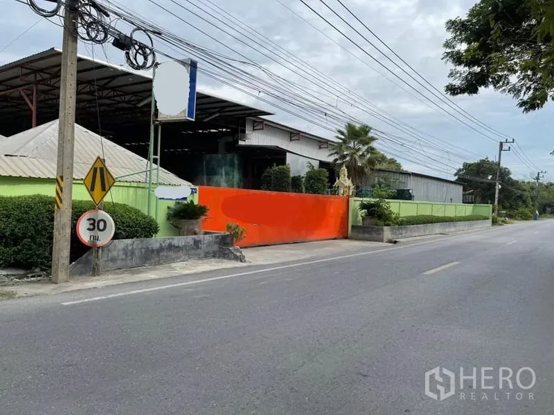 Warehouse for sale in Lam Luk Ka, Pathum Thani - Angled view of the warehouse frontage showing an orange sliding gate, roadside signage, and a wide steel canopy.