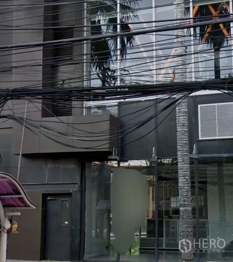 Retail Space for rent in Watthana, Bangkok - Street-level view of a modern dark-glass retail façade with full-height windows, a palm tree and overhead utility cables.