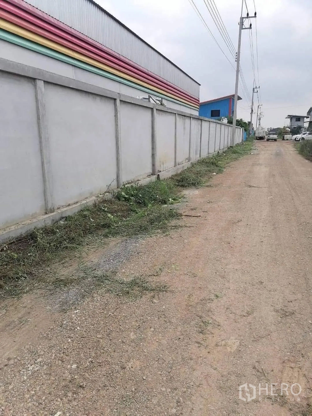 Warehouse for sale in Lam Luk Ka, Pathum Thani - Gravel frontage road running along a concrete perimeter wall and power poles next to the warehouse.