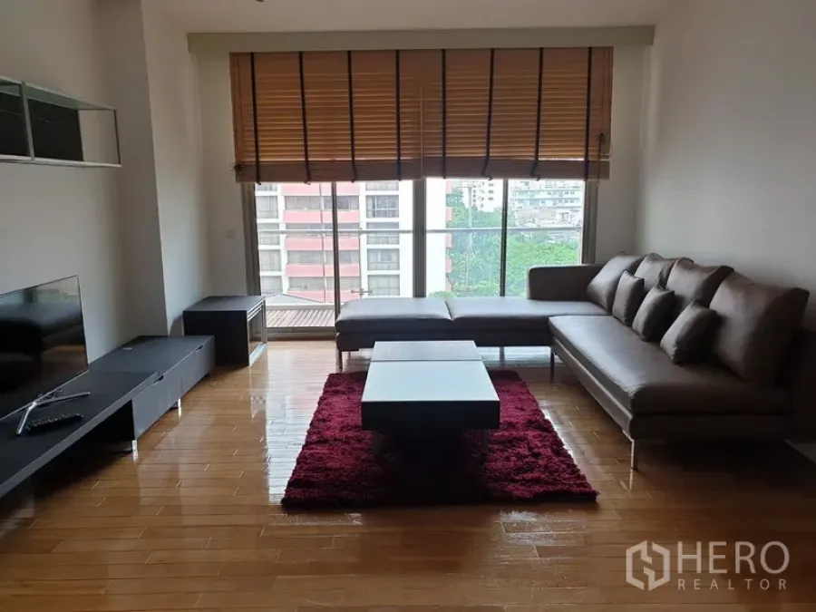 Condominium for rent in Bang Rak, Bangkok - Living room with L‑shaped sofa, TV console and coffee table on a red rug facing large windows with city view and balcony.