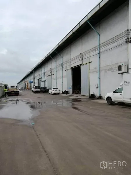 Warehouse for rent in Samut Prakan - Long row of industrial warehouses with truck loading area and parked vehicles on a wet concrete road.