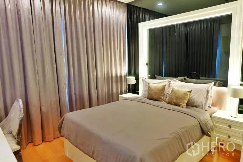 Condominium for rent in Khlong Toei, Bangkok - Comfortable bedroom with grey bedding, oversized illuminated mirror and blackout drapes.