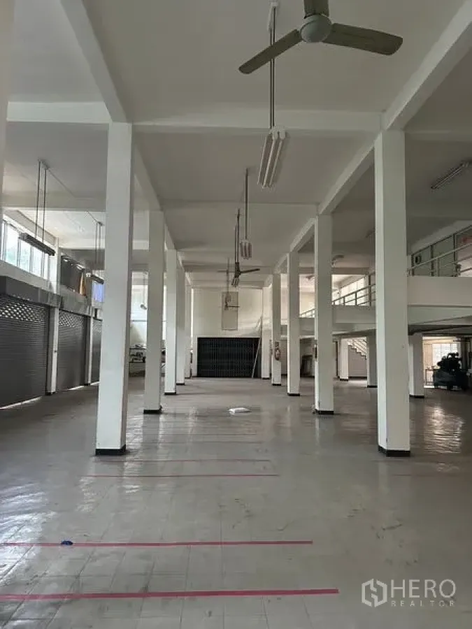 Shophouse for rent in Khlong Toei, Bangkok - Spacious shophouse interior with high ceiling, tall columns, ceiling fans, fluorescent lights, tiled floor and multiple roller shutters.
