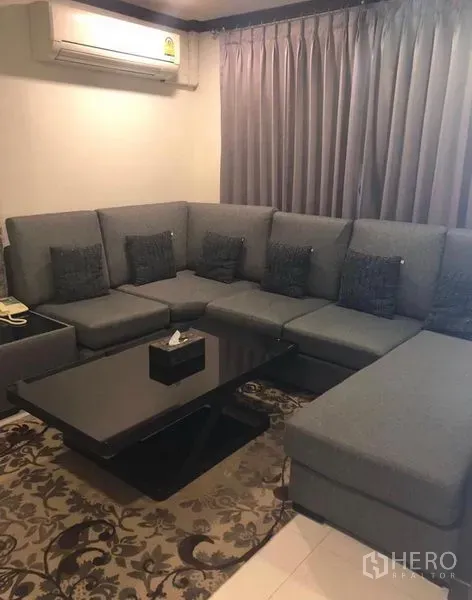 Townhouse for rent in Khlong Toei, Bangkok - Cozy living room featuring a large gray sectional sofa, blackout curtains, air conditioner, and a black coffee table.