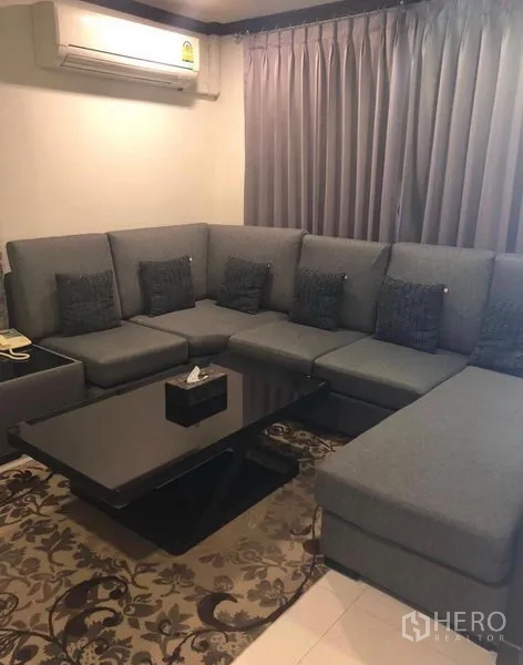 Townhouse for rent in Khlong Toei, Bangkok - Cozy living room featuring a large gray sectional sofa, blackout curtains, air conditioner, and a black coffee table.
