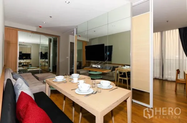 Condominium for rent in Khlong Toei, Bangkok - Dining area with bench seating facing a wall-mounted TV and mirrored wall, with floor-to-ceiling curtains at the window.