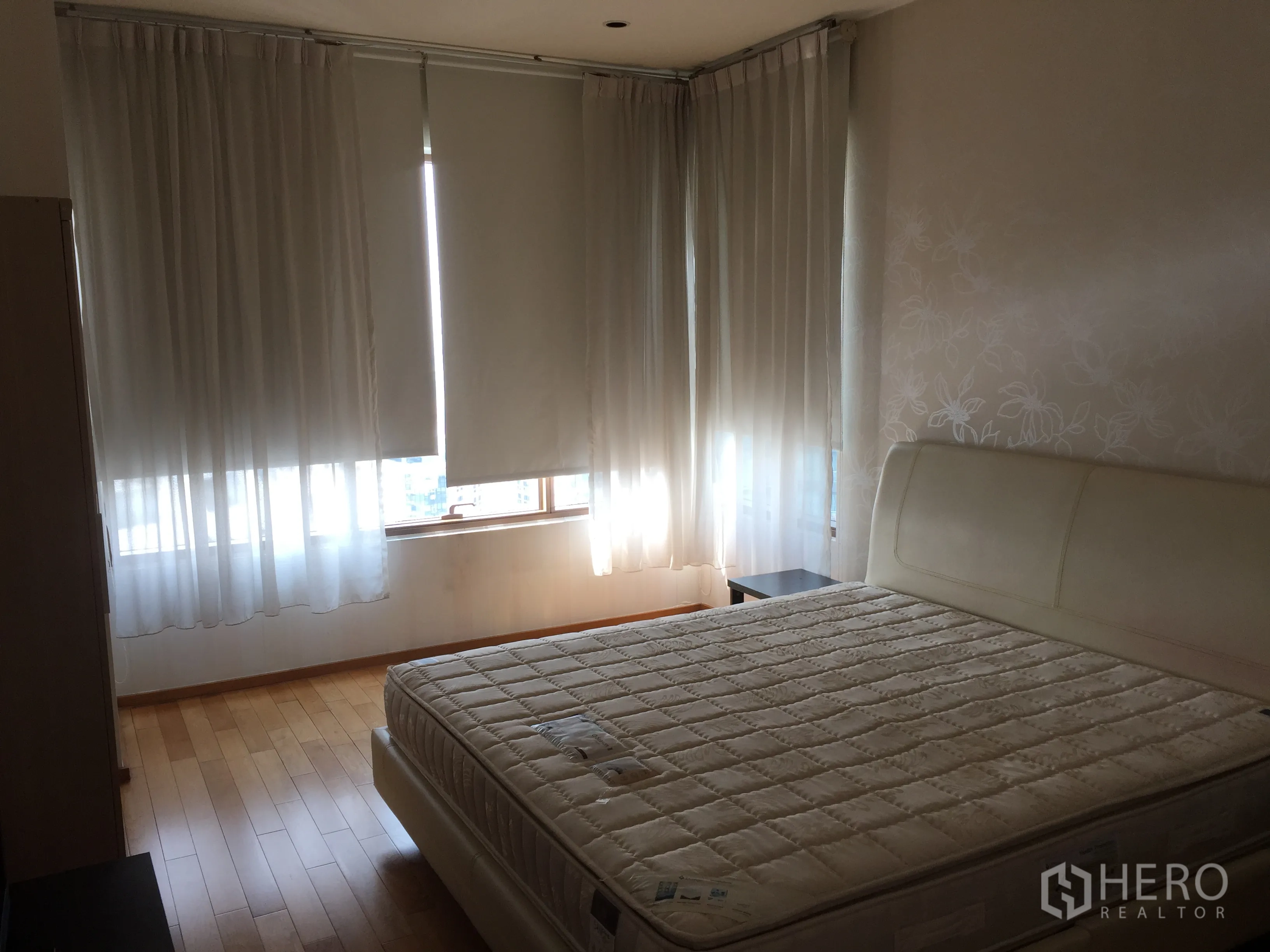 Condominium for rent or sale in Khlong Toei, Bangkok - Master bedroom with king-size bed, wood flooring, and wraparound windows with blinds and sheer curtains.