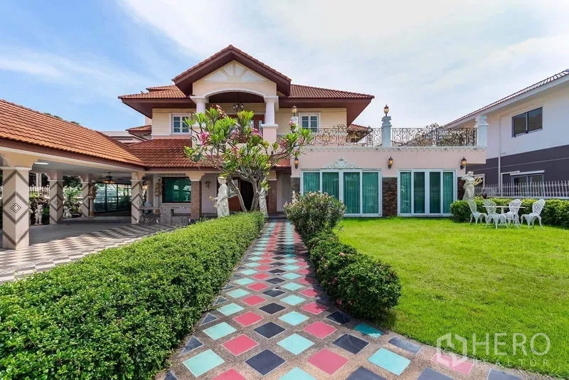 Detached House for rent or sale in Bang Khen, Bangkok - Colorful tiled walkway leading to a two‑storey detached house with red tile roof, balcony, statues, and a large manicured lawn.
