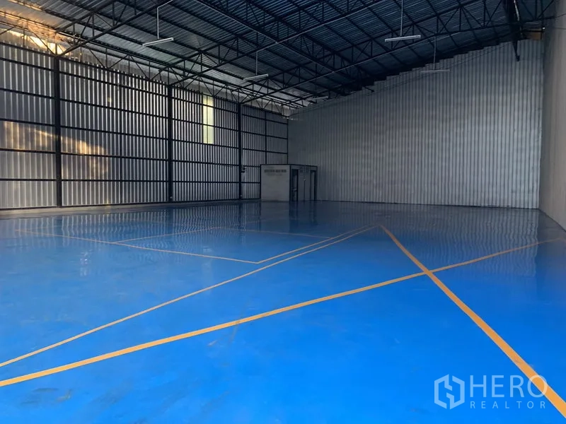 Warehouse for rent in Khan Na Yao, Bangkok - Spacious warehouse interior with a high steel truss ceiling and blue epoxy floor marked with safety lines.