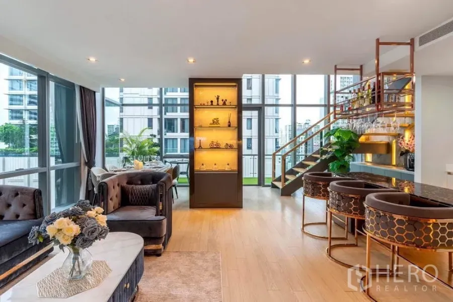 Condominium for rent in Watthana, Bangkok - Bright duplex living area with floor-to-ceiling windows, display cabinet, lounge seating and staircase beside a breakfast bar.