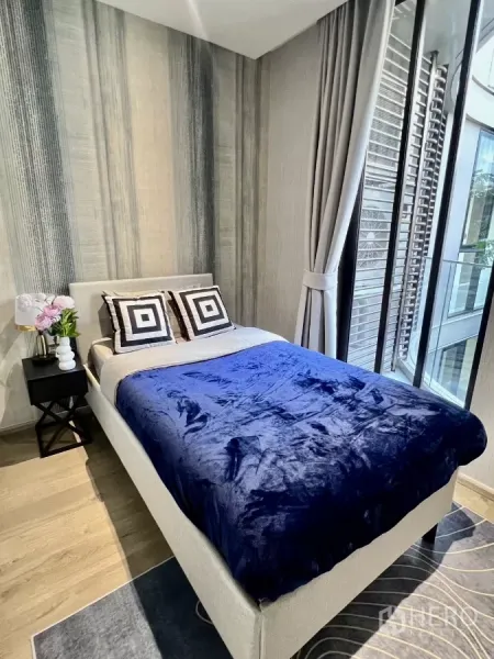 Condominium for rent in Khlong Toei, Bangkok - Cozy bedroom with blue throw and floor-to-ceiling windows overlooking the courtyard.