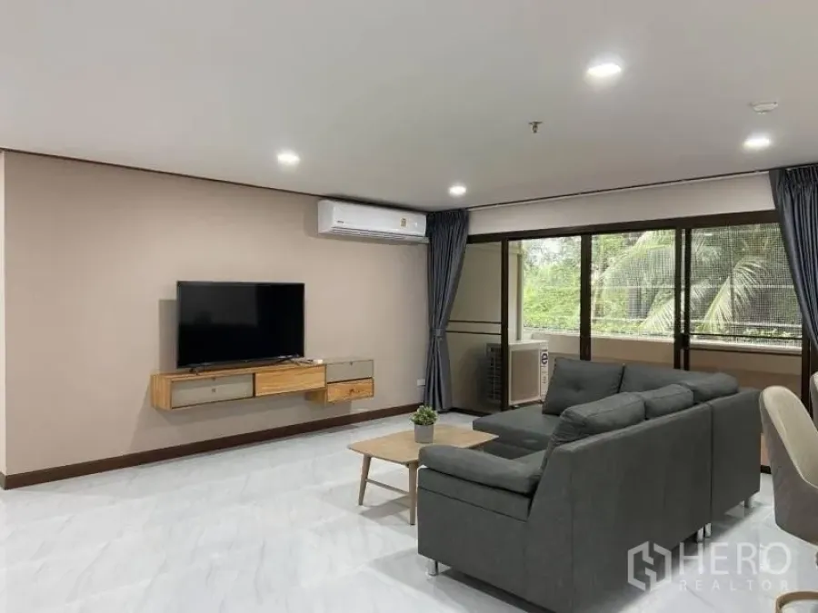 Condominium for rent or sale in Watthana, Bangkok - Open-plan living room with gray sectional sofa, wall-mounted TV and wide sliding doors to the balcony.