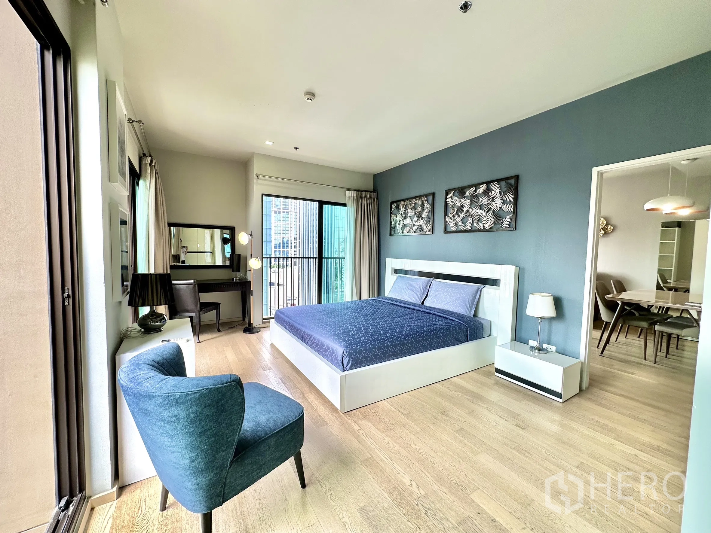 Condominium for rent in Bangkok - Spacious bedroom with king bed, blue accent wall, vanity desk and sliding doors to a balcony with city views.