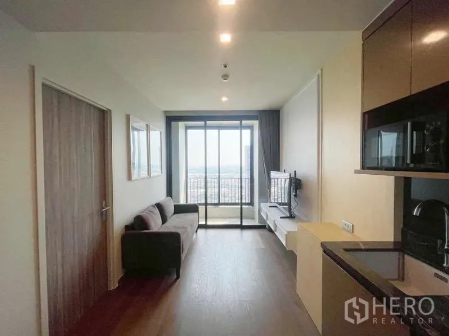 Condominium for rent in Khlong Toei, Bangkok - Bright living room with sofa, compact kitchenette and sliding glass doors opening to a balcony with city view.