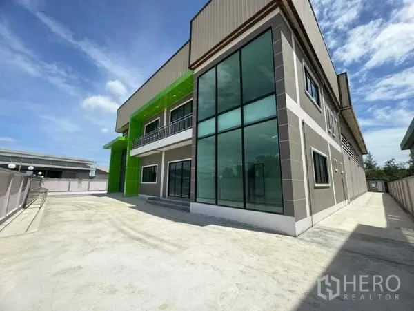 Factory for sale in Samut Sakhon - Side view of factory with tall glass corner windows and a wide concrete yard for parking.