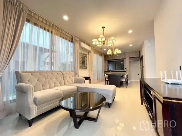 Detached House for sale in Bang Kapi, Bangkok - Bright living room with L-shaped tufted sofa, round coffee table, chandeliers and floor-to-ceiling curtains.