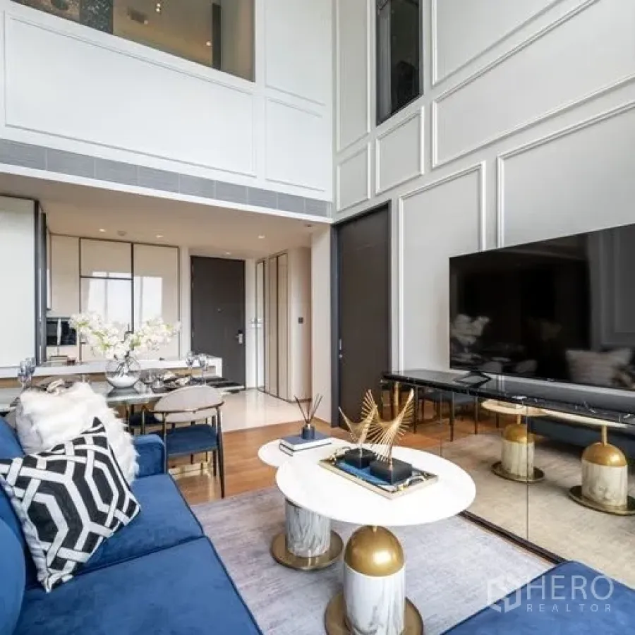 Condominium for rent in Khlong Toei, Bangkok - Contemporary living room featuring a blue sectional, nesting coffee tables, large TV console and tall paneled walls beside floor-to-ceiling windows.