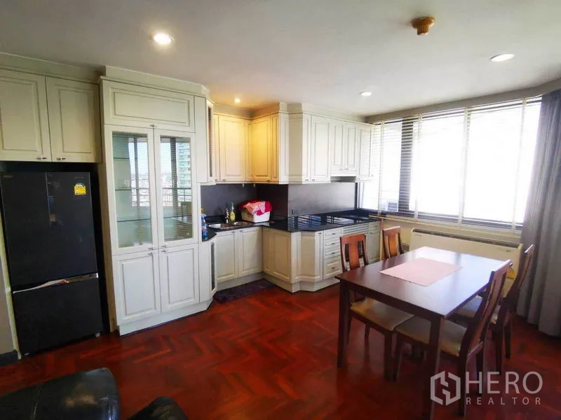 Condominium for rent in Khlong Toei, Bangkok - Sunny kitchen with cream cabinetry, black countertops and dining table by wide windows.