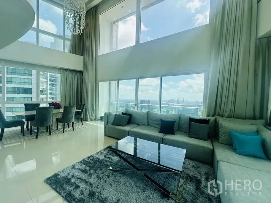 Condominium for rent or sale in Khlong Toei, Bangkok - Duplex penthouse living room at Millennium Residence with double-height ceiling, floor-to-ceiling windows, sofa set and dining area overlooking Benjakitti Park and Bangkok skyline.